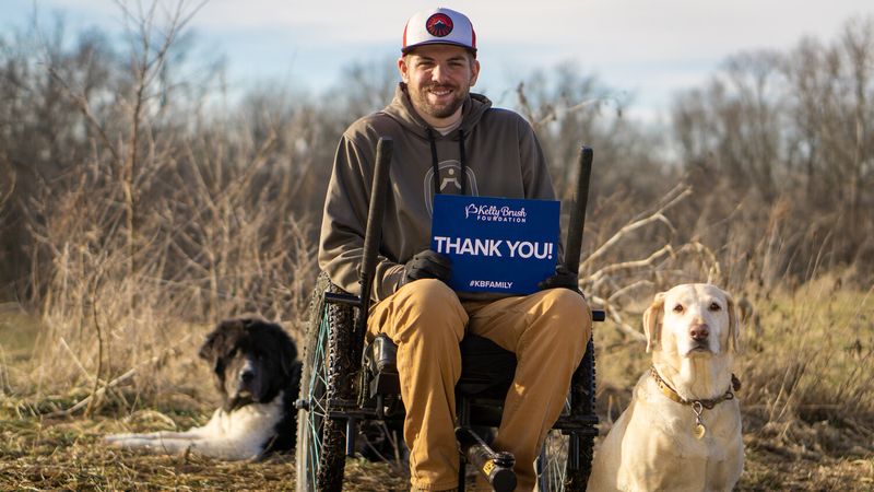 Adaptive Hunting with Ty Hockett: person holds Kelly Brush Foundation “thank you!” sign in GRIT Freedom Chair with two dogs near bare bushes
