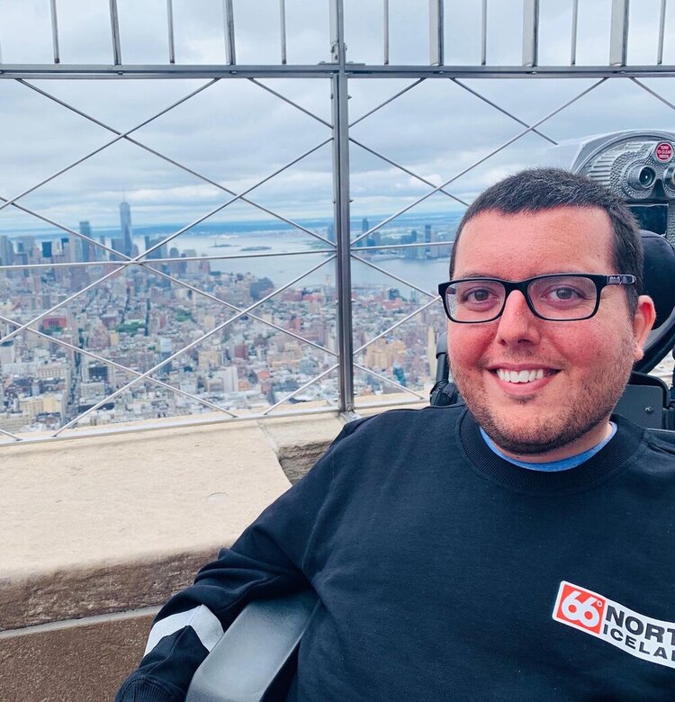 Accessible Hiking Trails You’ve Never Heard Of, author Cory Lee smiles with rooftop view of city and water behind him in his wheelchair