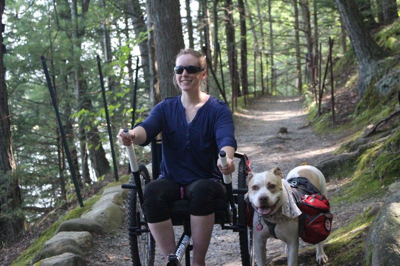 Flying With the GRIT Freedom Chair rider perspective: Nerissa Cannon rides GRIT outdoor wheelchair on bumpy dirt path through trees with dog