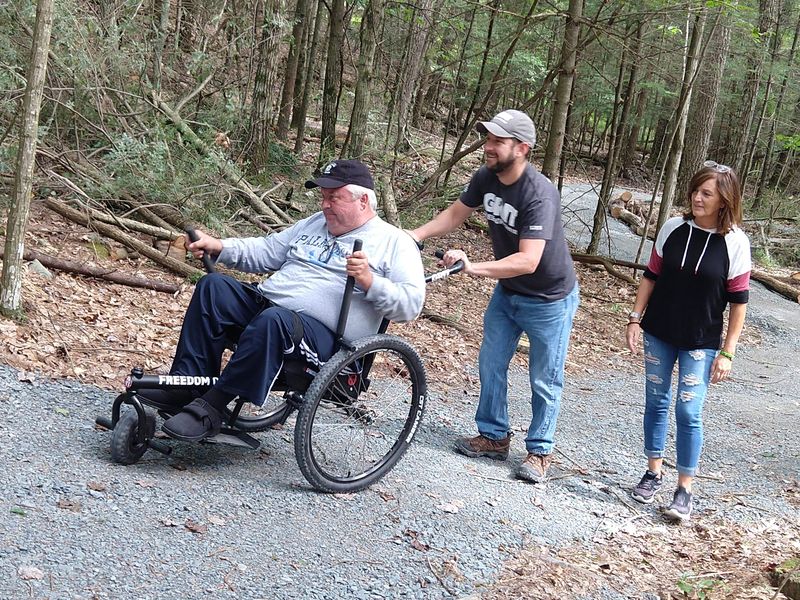 How To Ask for Personal Mobility Equipment Funding Help Step #3, Ask: 3 adults hike uphill on gravel, 1 in GRIT Freedom Chair with 1 pushing
