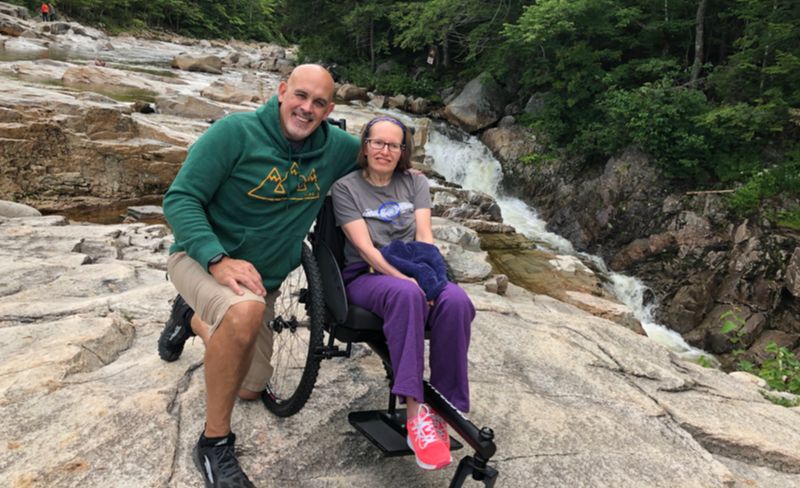 Patrick kneels next to Colleen in GRIT Freedom Chair all terrain wheelchair on large rocks next to running water