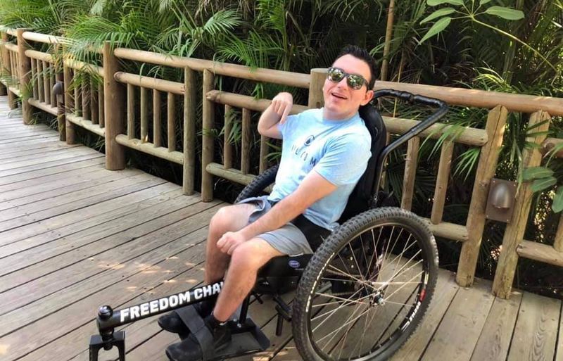 Trandon Mechling: Spartan, GRIT Athlete: Trandon smiles using his GRIT Freedom Chair outdoor wheelchair on wooden bridge by tropical trees